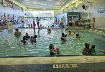 In the activity pool at the Bear Levin Studer YMCA a group of parents and young children participate in a Swim Starters class, while other swimmers enjoy the lap pool in the background.