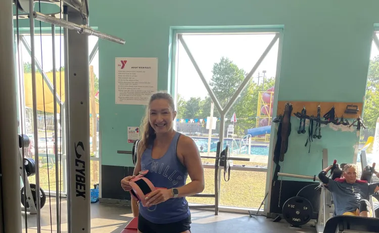Group Exercise Instructor and Personal Trainer Anastasia smiles for a photo on the wellness floor in front of the windows looking out to the pool.