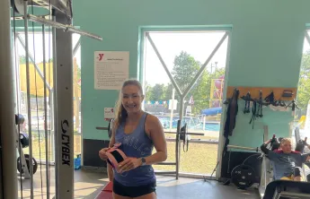 Group Exercise Instructor and Personal Trainer Anastasia smiles for a photo on the wellness floor in front of the windows looking out to the pool.
